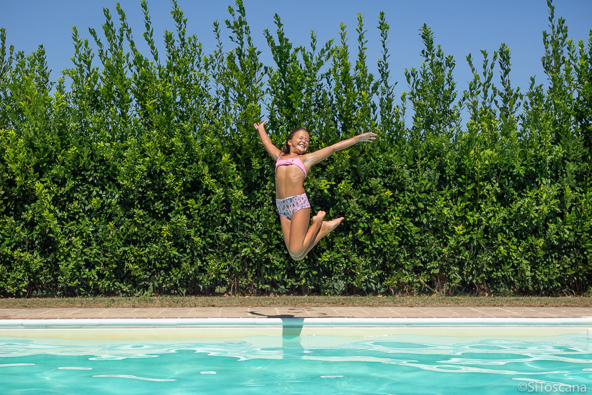 Bildet viser ei jente som hopper ut i et basseng i Toscana. Det er sommer. Jenta er glad og smiler.
