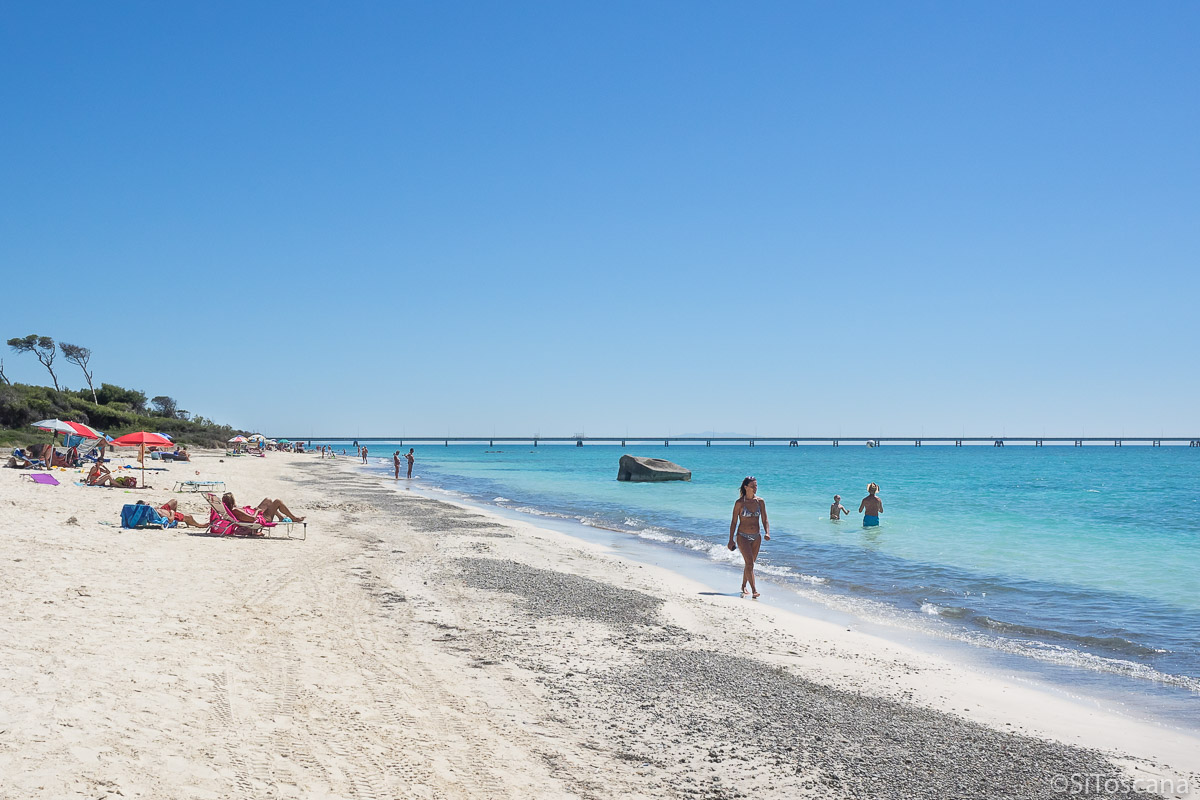 Bildet viser en kvinne som går på en strand i Toscana. det er sommer. Vannet er turkisblått.