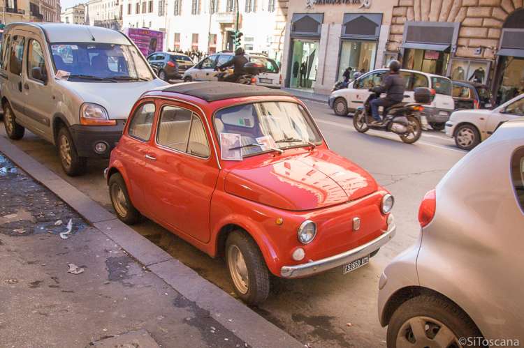 Bildet viser en gammel rød Fiat 500 parkert i ei bygate. Andre biler er parkert foran og bak. I gata kjører bilder og scootere forbi.