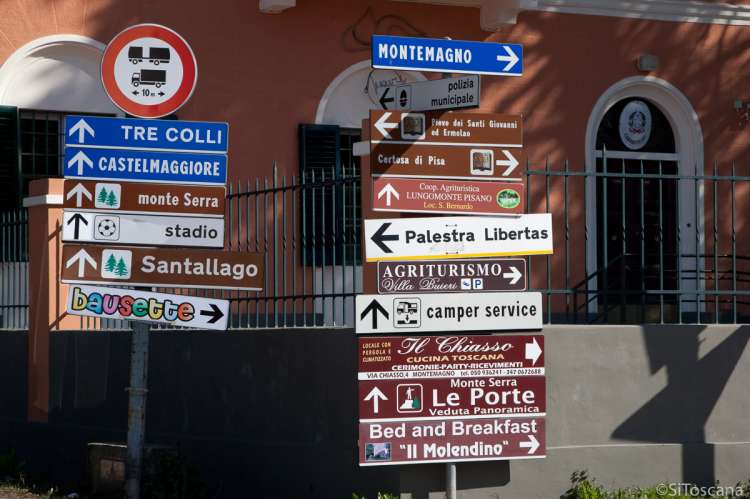 Bildet viser trafikkskilt og informasjonsskilt i et gatekryss i Calci, Toscana, Italia. Det er en blanding av trafikkskilt, opplysningsskilt og reklameskilt.