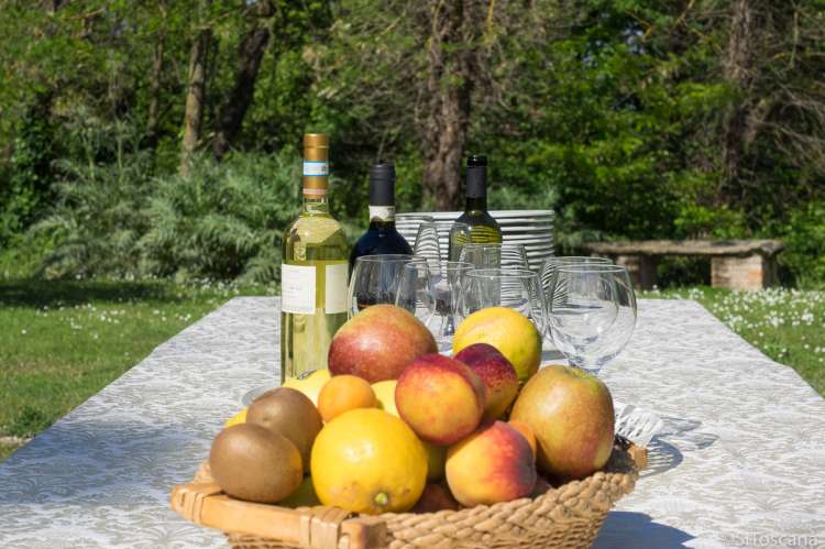Langbord klart for oppdekking utenfor et av SiToscana sine feriehus. Det er en vakker maidag med sol og blomster i gresset. På bordet står det fat, vinglass, vinflasker og frukt.