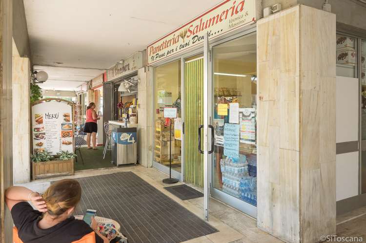 Bildet viser inngangen og vinduet til en Panetteria og Salumeria i badebyen Tirrenia i Toscana. I forgrunnen sitter en kvinne på en stol utenfor og ser på telefonen sin. I bakgrunnen står en kvinne og ser innen på et spisested som ligger vegg i vegg.