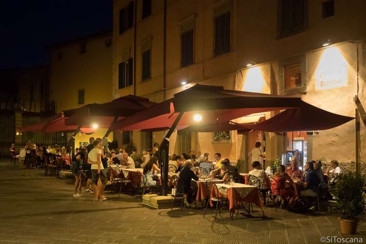 Bildet viser folk som sitter ute og spiser på en av restaurantene på Piazza Dante Alighieri i Pisa. Bildet er tatt en varm sommerkveld og det er mørkt.