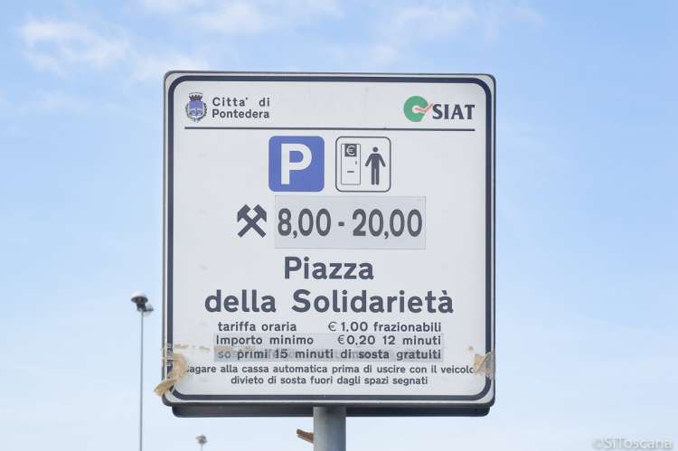 Bildet viser et parkeringsskilt på den store parkeringsplassen Piazza della Solidarietà i byen Pontedera. På skiltet står åpningstider, takster og at du må betale i cassa automatica/betalingsautomaten før du kjører ut.