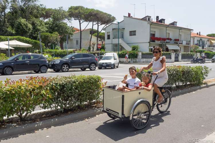 Bildet viser en kvinne som sykler på en av de oppmerkede sykkelbanene i Tirrenia. Sykkelen har en vogn foran hvor det sitter en gutt og ei jente og spiser is.