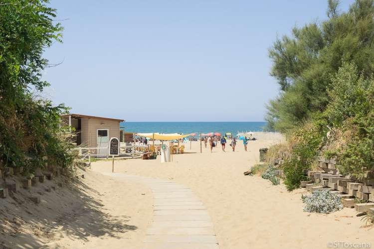 Bildet viser nedgangen til stranda Spiaggia Verruca i badebyen Tirrenia i Toscana. Du ser huset til et spisested med terrasse, og mennesker som er på vei opp fra stranda. I det fjerne skimtes Middelhavet med krystallblått hav. Det er sol og blå himmel.