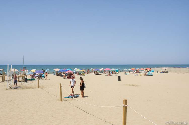 Bildet viser spiaggia libera på Spiaggia Verruca i badebyen Tirrenia. Du ser dusjfasilitetene, et tau som skiller fristranda fra der du kan leie parasoll etc. Folk som ligger under medbrakte parasoller. Og i bakgrunnen strandkanten og Middelhavet.