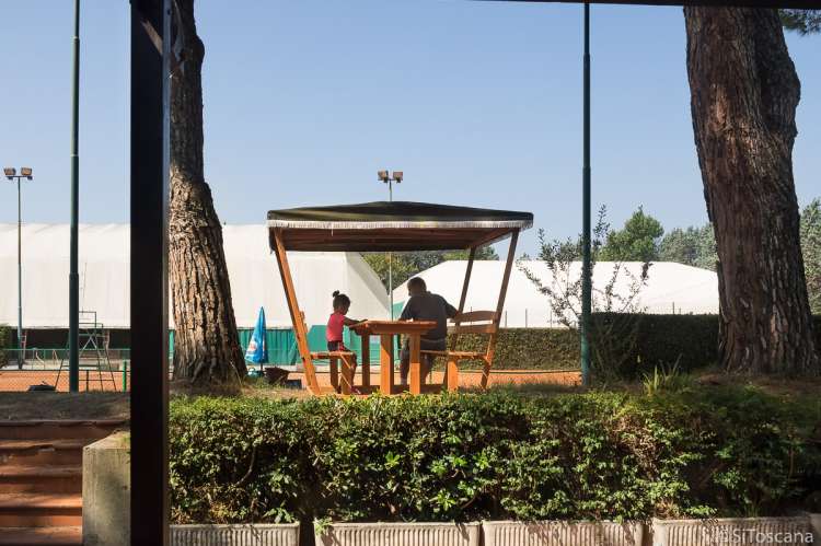 Bildet viser en mann og ei jente som sitter ved et bord under en pergola og prater. Ved tennisbanene i La Gabella ved Calci, Pisa.