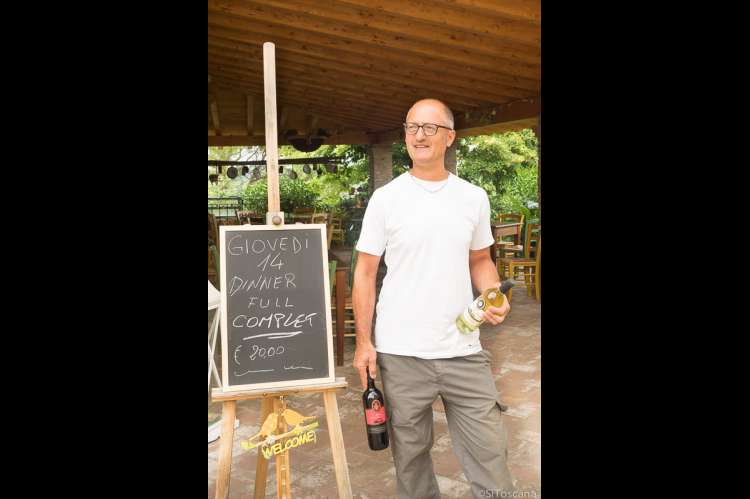 Verten Lamberto reklamerer for flere retters middag til 20 euro, inkludert vin! Foto av vertskapet ved vinbonden foran en plakat der restauranten på vingården inviteter til et italiensk middagsmåltid for 20 euro.