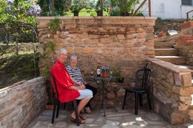 Uteplass ved inngangspartiet. Hagen er ovenfor. Bilde av to ferierende damer på terrassen ved inngangspartiet. Steinmurer, vin og blomster.