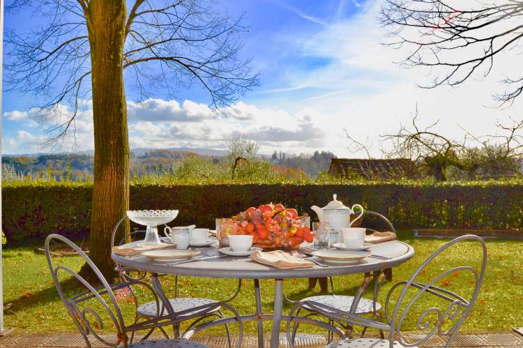 Bildet viser en av terrassene i Villa Alba. Et bord er dekket for kaffe og kaker. Utsikt mot åsene rundt. Det er sol og grønt.