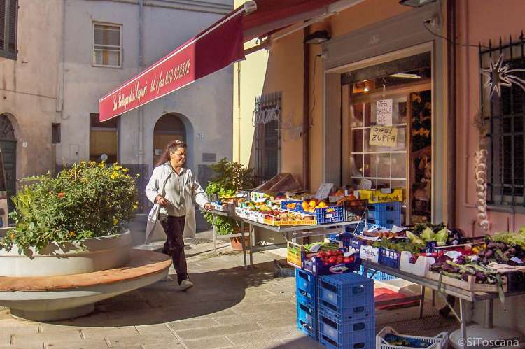 Bildet viser en kvinne utenfor en av nærbutikkene i Calci. Frukt og grønnsaker ligger utstilt  på bord.