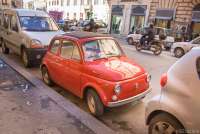 Bildet viser en gammel rød Fiat 500 parkert i ei bygate. Andre biler er parkert foran og bak. I gata kjører bilder og scootere forbi.