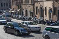 Bildet viser biler som kjører i en travel bygate. I bakgrunnen står mange scootere parkert på en scooterparkering.