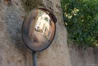 Bildet viser et sladrespeil foran en murvegg i middelalderbyen Montemagno ved Pisa. Du ser en skarp sving i ei smal gate, husvegger og en bil som kjører. Speilet brukes til å se trafikken som kommer i mot.