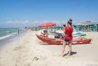 Bildet viser Karine som jobber for SiToscana på en av strendene i Tirrenia. Rød redningsbåt foran badevakter under rød parasoll. Og mange mennesker som bader. Det er sol og sommer.