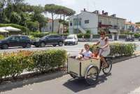 Bildet viser en kvinne som sykler på en av de oppmerkede sykkelbanene i Tirrenia. Sykkelen har en vogn foran hvor det sitter en gutt og ei jente og spiser is.