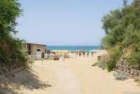 Bildet viser nedgangen til stranda Spiaggia Verruca i badebyen Tirrenia i Toscana. Du ser huset til et spisested med terrasse, og mennesker som er på vei opp fra stranda. I det fjerne skimtes Middelhavet med krystallblått hav. Det er sol og blå himmel.