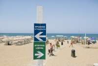 Bildet viser et skilt med teksten Spiaggia attrezzata og Spiaggia libera på Spiaggia Verruca i Tirrenia i Toscana. I bakgrunnen skimtes mennesker på vei opp fra stranda, parasoller og solstoler samt stranda og Middelhavet.