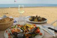 Bildet viser sjømat på tallerken, vinglass med hvitvin og utsikt til sandstrand og hav. Vicino Mare ved kysten sør i Toscana.